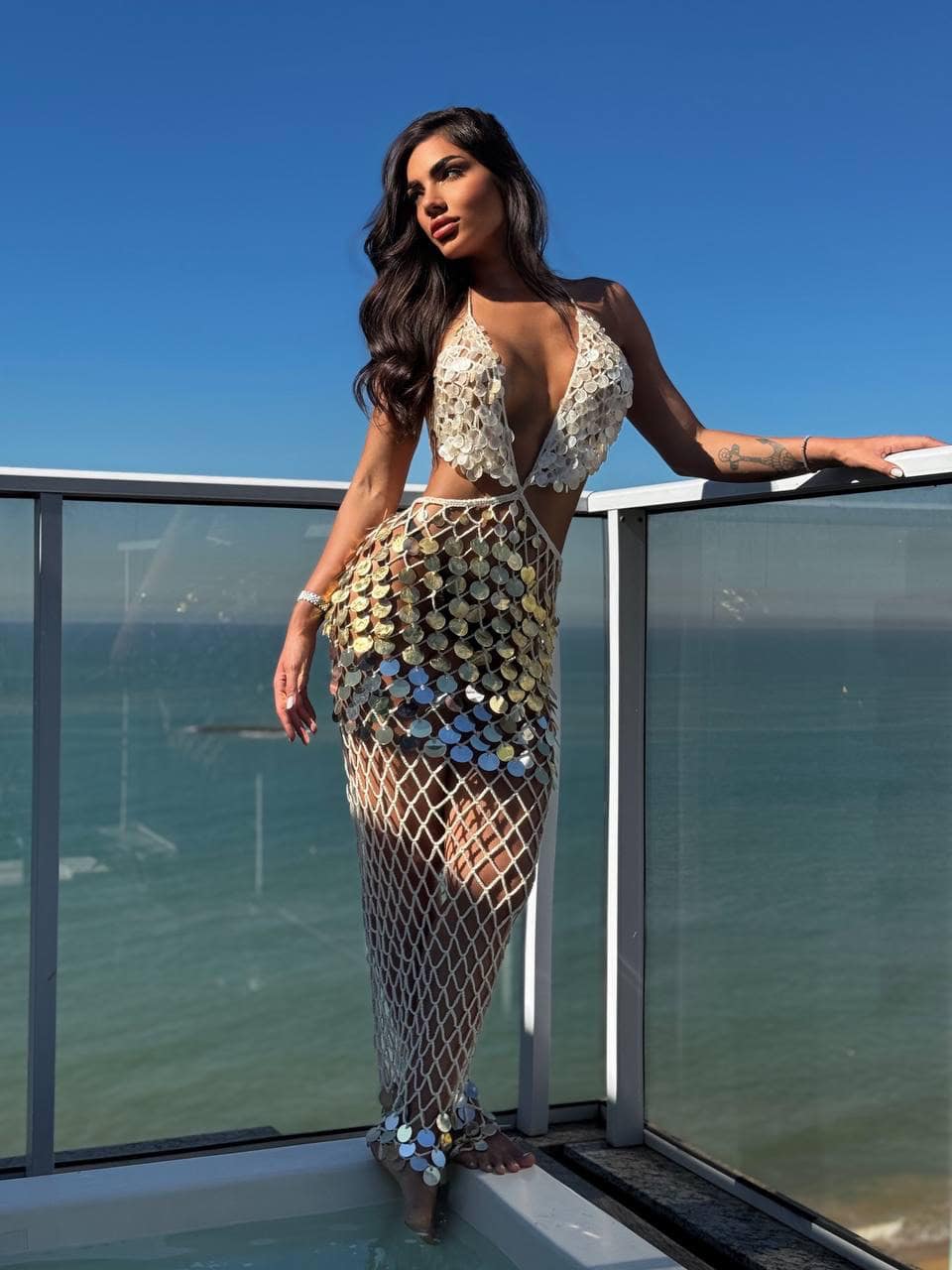 Woman in shimmering crochet dress on balcony overlooking the sea.