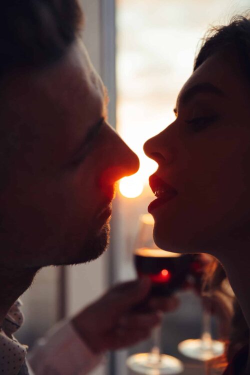 portrait of lovers kissing at sunset and holding glass with wine at home