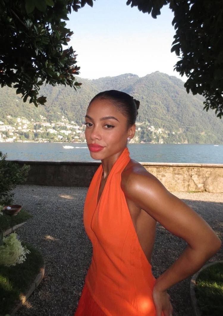 Woman in an orange halter dress posing before a lake.