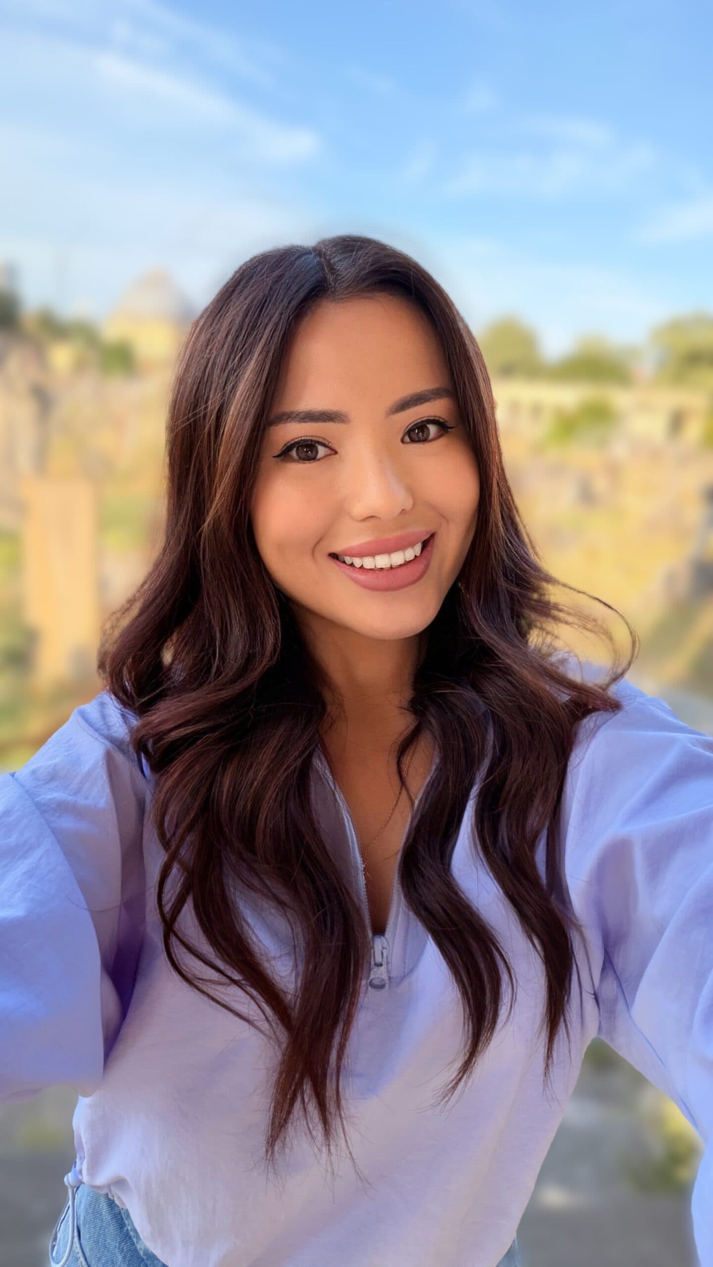 Smiling woman with wavy brown hair posing outdoors near ruins.